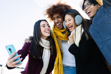 Diverse group of teenage friends having fun taking selfie portrait at city street on winter. Holiday lifestyle and technology concept