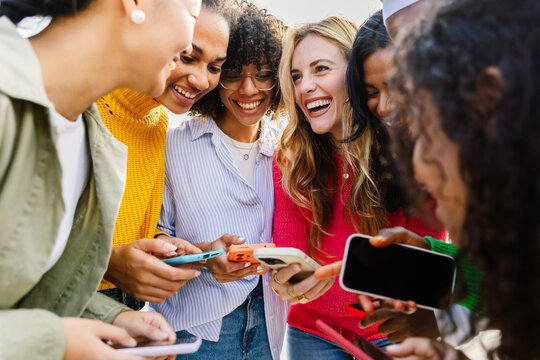 Happy diverse women looking at smartphone screen and smiling outdoors. Multiracial female friends standing together enjoying social media interaction. Technology and connection concept. - Powered by Adobe