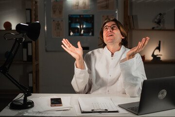 Angry General practitioner doctor sitting at desk working on night shift makes omg gesture with...