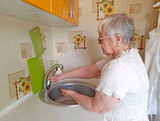 An elderly woman wearing glasses washes her hands or an object in a metal sink in the kitchen.