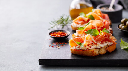 Crispy bruschetta topped with jamon, creamy cheese and fresh basil leaves. Grey background. Close up. Copy space.