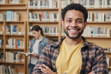 Portrait of young adult Black man smiling with arms crossed in library, standing in foreground while Caucasian young adult woman reading book in background near bookshelves