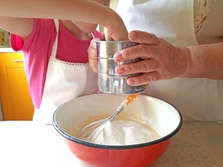 A grandmother and a child sift flour through a metal sieve.