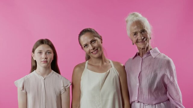 Studio portrait shot of smiling adult woman posing with elderly mother teenage daughter holding hands on shoulders then turning to camera against pink background, slow motion