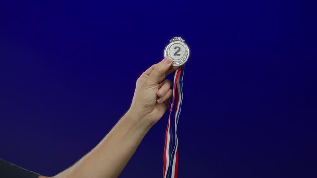 Man hand holds a silver medal with ribbons against a plain blue background, symbolizing achievement and second place victory.