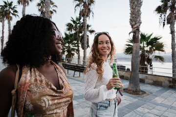 Diverse young women friends walking seaside with beer