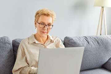 Adult woman wearing glasses sitting on comfortable gray sofa, focused on working from home on her laptop. Productive busy female remote employee making video call, writing message, solving work tasks.