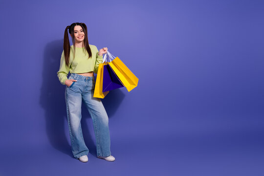 Trendy young woman holding bright shopping bags in a stylish outfit against a vibrant purple backdrop