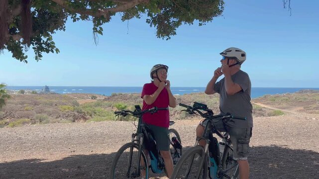 Caucasian senior couple of cyclists wearing helmets enjoying healthy lifestyle riding with electric bicycles in a sunny day. Authentic senior retired life concept. Horizon over the sea