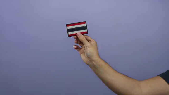 Caucasian man holding embroidered thailand flag patch against a purple wall background.