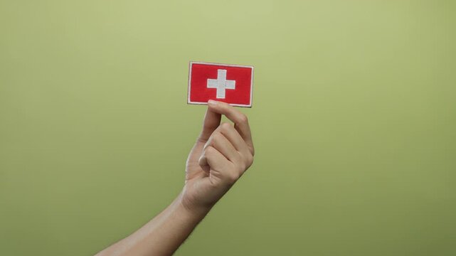 Caucasian man holding swiss flag emblem with embroidery against an isolated yellow wall background, highlighting national identity and cultural expression.