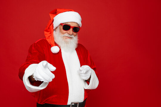 Close up portrait of funny happy excited old bearded Santa Claus face wearing hat, glasses, looking at camera, showing pointing finger gesture choosing you for advertising isolated on red background.