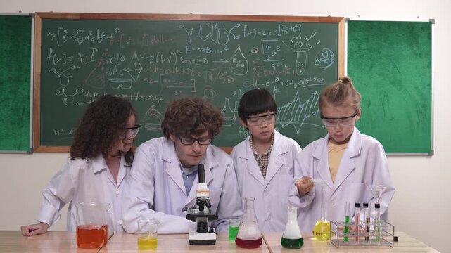 Smart caucasian teacher looking under microscope while diverse children doing experiment at STEM science lesson. Professional happy scientist inspect biochemical example by using microscope. Pedagogy.