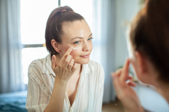 Adult woman applying eye cream at home mirror, calm and content