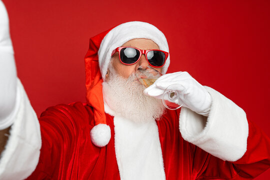 Selfie shot of retired old man grey beard funky holding drinking full glass golden wine drink having fun wear santa x-mas costume suspender sunglass cap isolated red color background