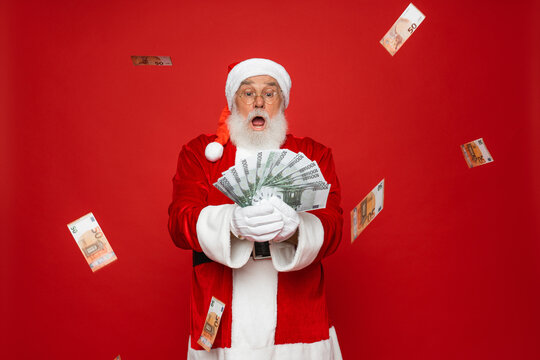 Photo of pensioner old man grey beard raise fists open mouth money shower hold cash wear Santa Claus traditional costume red coat hat standing isolated red background