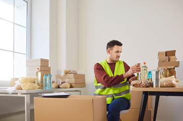 Male volunteer in reflective vest sorting and packing food supplies on tables in food bank. Humanitarian aid worker preparing donation boxes with essential items for charity and community support.