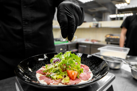 Professional chef plating gourmet beef carpaccio appetizer in a restaurant kitchen with fresh ingredients.
