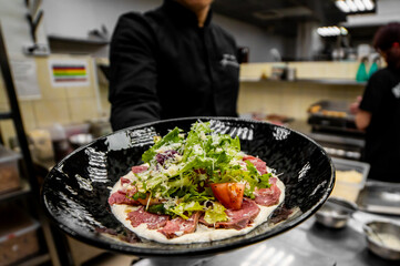 A close-up, low-angle photo of beef carpaccio with lettuce, parmesan cheese, and tomato on a black plate, served by a person in a restaurant kitchen