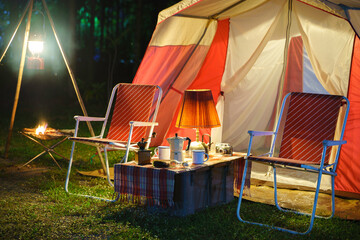 Camping tents at night, vintage cabin tents, antique oil lamps, retro chairs, and a group of camping tents with outdoor coffee-making facilities on wooden tables in a forest camping area.