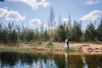 Couple in wedding attire stands by serene lake, surrounded by lush trees and blue sky, capturing a romantic moment in nature's tranquil setting