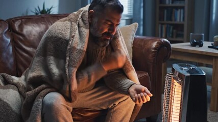 Man wrapped in a blanket, feeling cold, sitting near a heater in a living room