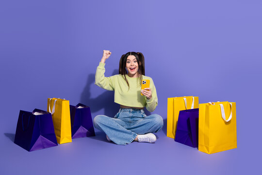 Young woman sitting on colorful background holding smartphone celebrating surrounded by shopping bags while smiling