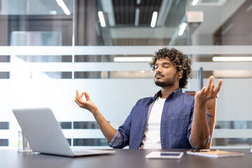 Relaxed and calm young Indian man sitting in an office chair at a desk in a lotus position and meditating