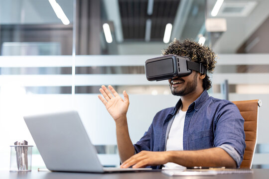 Young Indian man in a virtual mask sits in an office at a table with a laptop, gesturing with his hands - Powered by Adobe