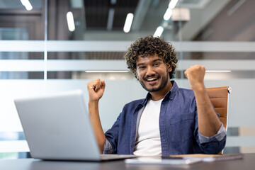 Portrait of a happy young Indian man sitting in the office at a table with a laptop, raising his hands up and rejoicing
