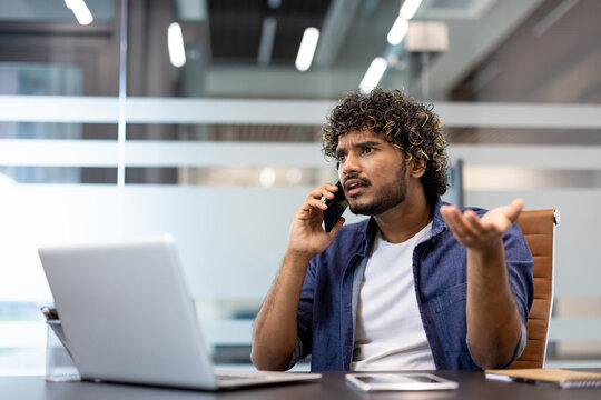 Worried young Indian man sitting at a desk in the office, talking on the phone and spreading his hands in frustration - Powered by Adobe