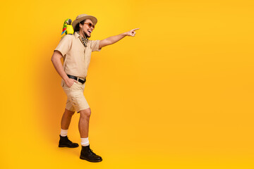 Man in safari outfit points across a bright yellow backdrop with parrot on his shoulder conveying...
