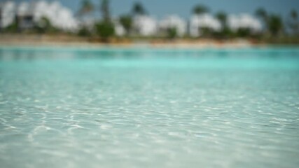 Glistening turquoise water gently ripples with a shallow depth of field, revealing a soft-focus view of a sandy beach, palm trees, and a luxurious resort hotel under a clear blue summer sky