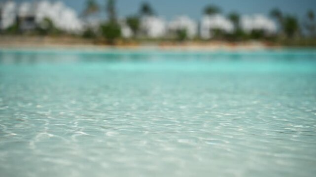 Glistening turquoise water gently ripples with a shallow depth of field, revealing a soft-focus view of a sandy beach, palm trees, and a luxurious resort hotel under a clear blue summer sky