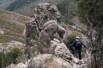 A male climber carefully descends a rocky section of a via ferrata in Castillo de Locub n, Ja n, Spain, wearing a helmet and safety gear amidst mountain scenery.