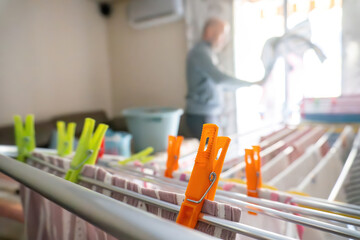Colorful laundry clips on a drying rack in sharp focus while a blurred adult man hangs clothes near a bright window. A natural domestic scene showing everyday household chores.