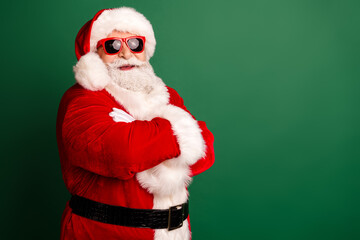 Santa in red velvet suit with white fur trim and hat stands with arms crossed against a green background for Christmas and holiday season