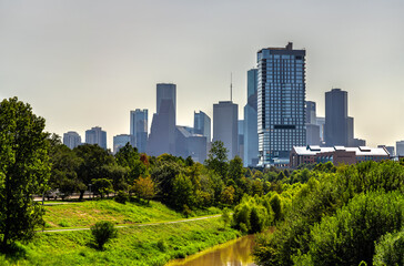Fototapeta premium Downtown Houston skyline rises above the lush green trees and path of Buffalo Bayou Park. The muddy brown bayou flows in the foreground