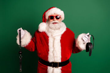Santa in red suit with white fur and shades holds leather straps against a green background ready for Christmas fun and holiday shopping promotion