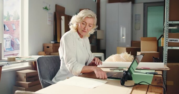 Writing, ecommerce and laptop with woman in warehouse for delivery confirmation, inventory and research. Shipping checklist, online order and courier distribution with mature person as entrepreneur