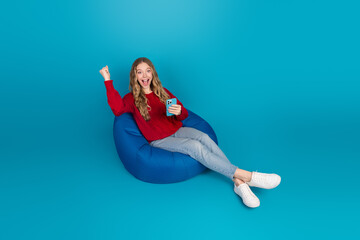 Cheerful young woman in casual outfit relaxing on a bean bag with a smartphone against a vibrant...
