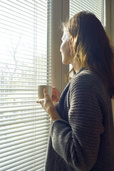 Woman holding a coffee mug and gazing through window blinds as morning sunlight illuminates her profile. Symbolizes peaceful morning ritual, contemplative solitude, and warm beverage comfort.