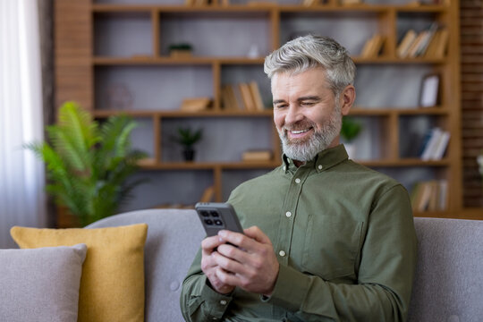 Smiling mature man with grey hair and beard sitting comfortably on a modern couch at home, engaging with his smartphone for communication, entertainment, or digital tasks