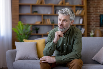 Mature man with gray hair and beard sitting on a comfortable sofa in a modern home interior, pensive and deliberating while looking away, reflecting on thoughts and plans