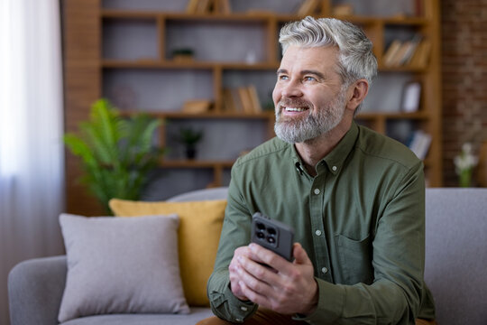 Mature man with grey hair and beard sitting on a couch in his modern living room, holding a smartphone and looking away with a happy, thoughtful expression, enjoying leisure time