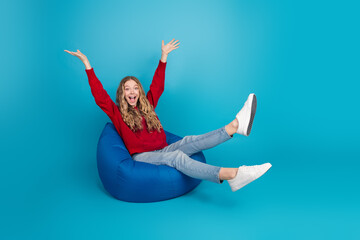 Adorable young woman in a red sweater sitting on a blue bean bag with a cheerful expression, full...