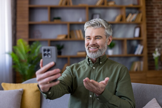Happy mature man with grey hair enjoying a video call on his smartphone, smiling and gesturing while communicating from the comfort of his home living room