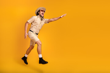 Adventure seeker in safari outfit leaps against bright orange backdrop smiling wearing hat and...