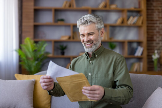 Mature man with gray hair and beard smiling brightly while opening a delivery envelope, reading a personal letter sitting comfortably on a sofa at home