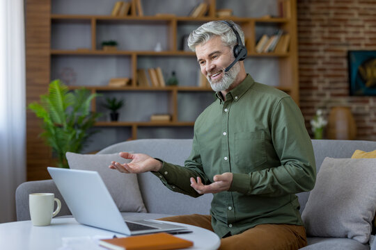 Mature man wearing a headset and using a laptop while sitting on a couch, actively participating in a video call or remote conference from his comfortable home environment